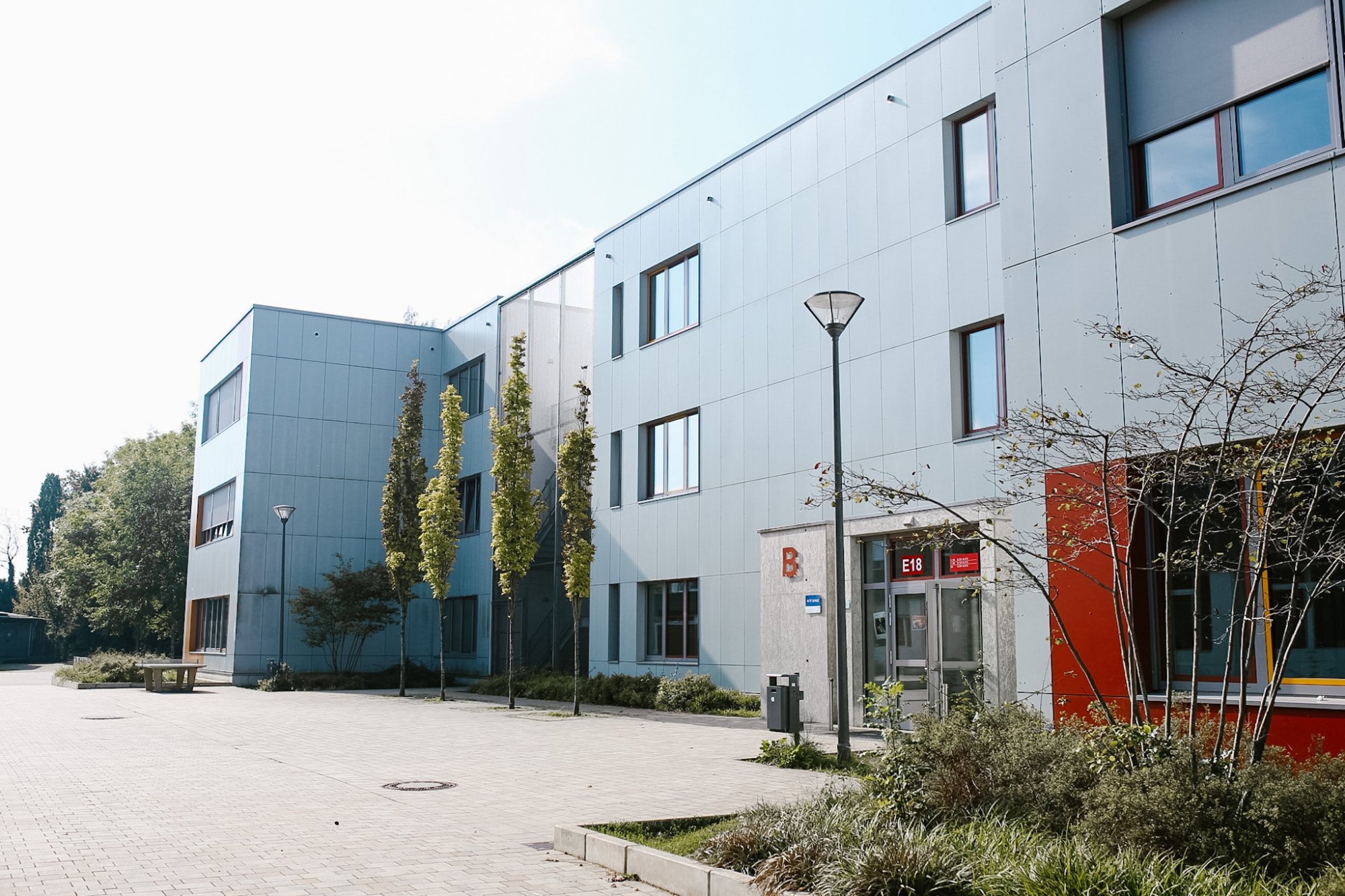 Gepflasterter Schulhof vor dem hellgrauen B-Gebäude mit Eingang E18. Am Weg stehen schmale junge Bäume, Laternen und eine Bank.