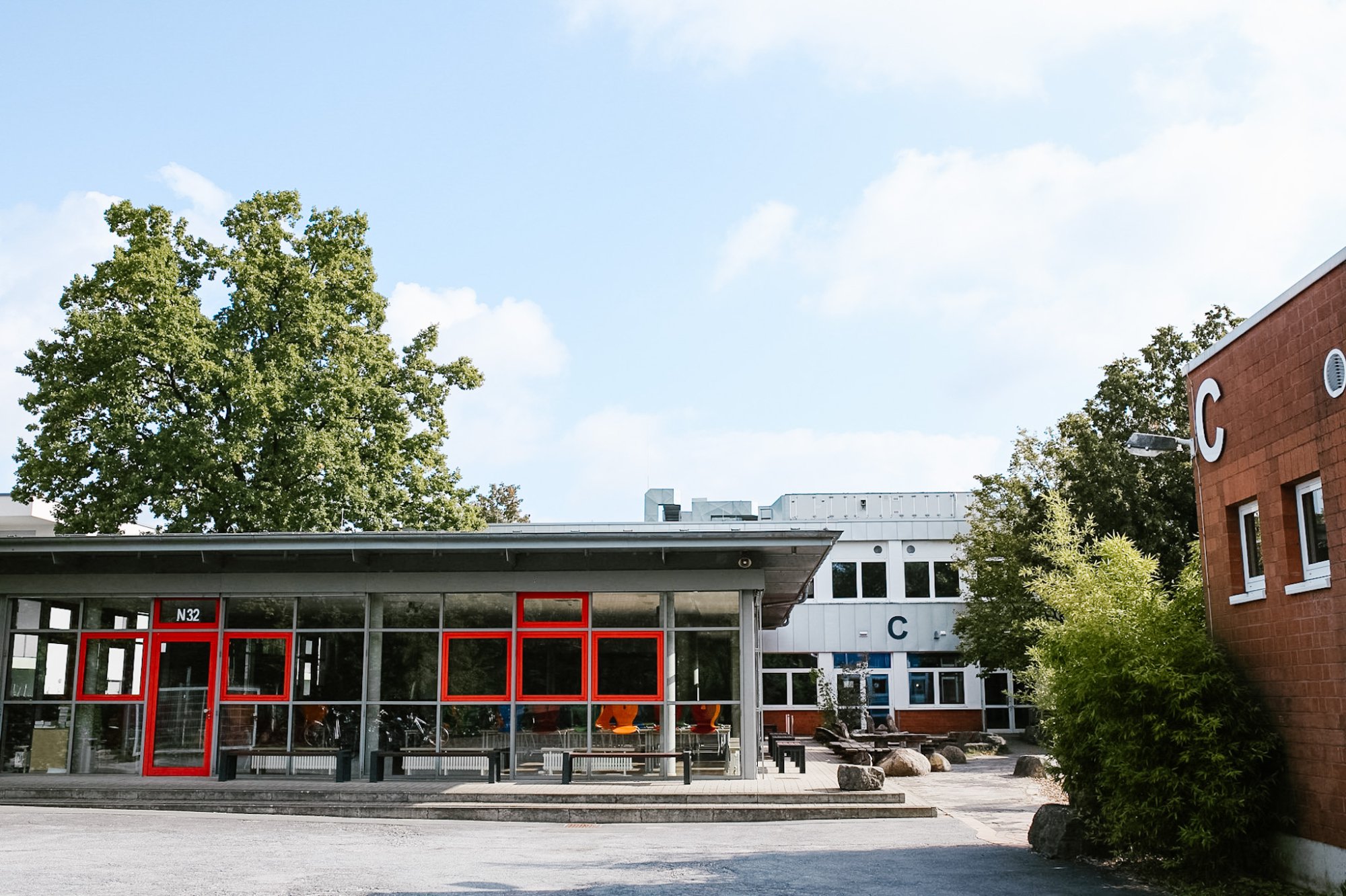 Schulhof mit verglastem Pavillon mit roten Fensterrahmen, Eingang N32. Im Hintergrund das C-Gebäude, rechts ein Klinkerbau mit einem großen C, große Bäume vor blauem Himmel.