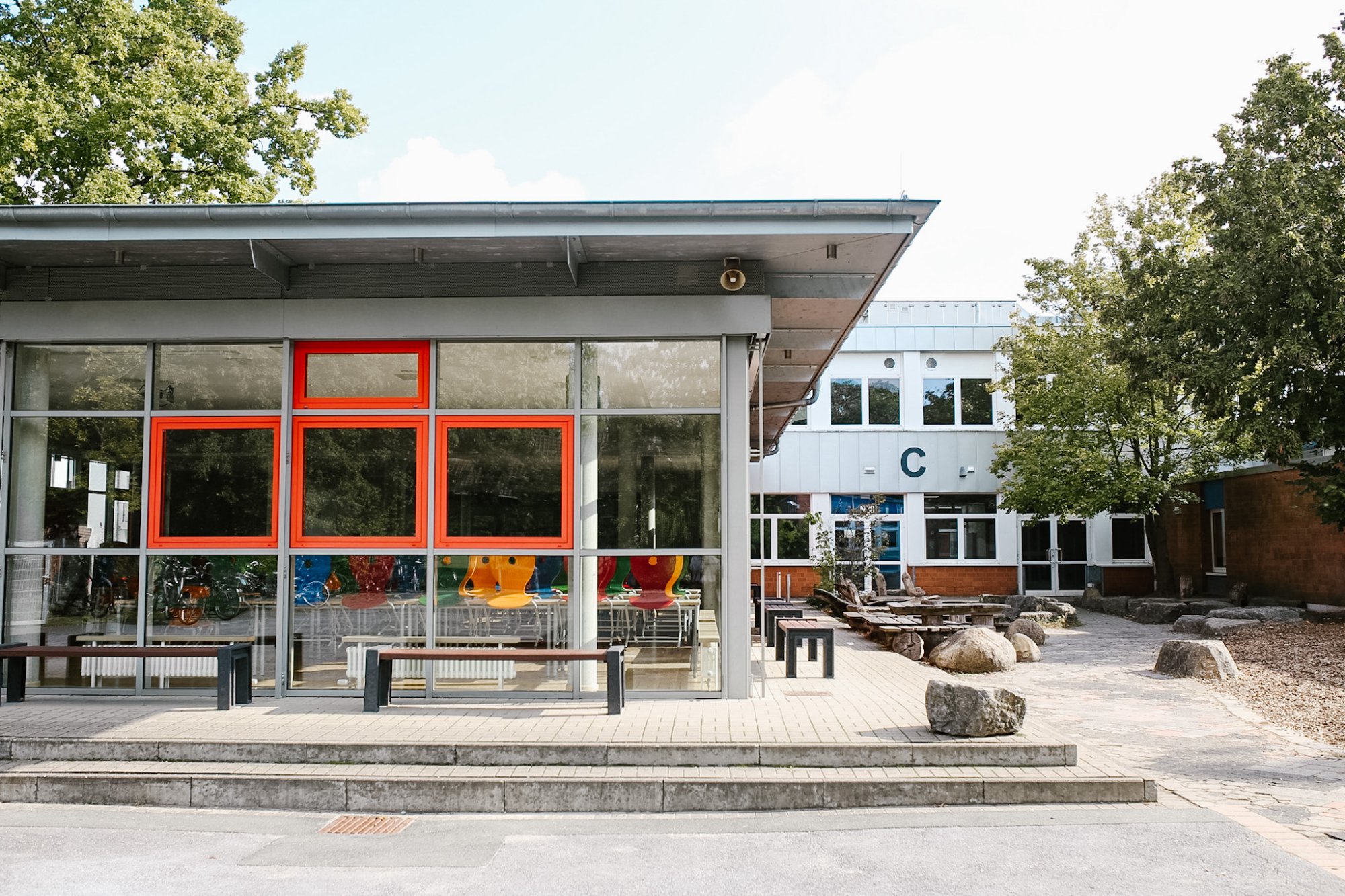 Verglaster Pavillon mit orangeroten Fensterrahmen und Bänken davor. Daneben ein Hofbereich mit Natursteinen und Holzbänken, dahinter das C-Gebäude.