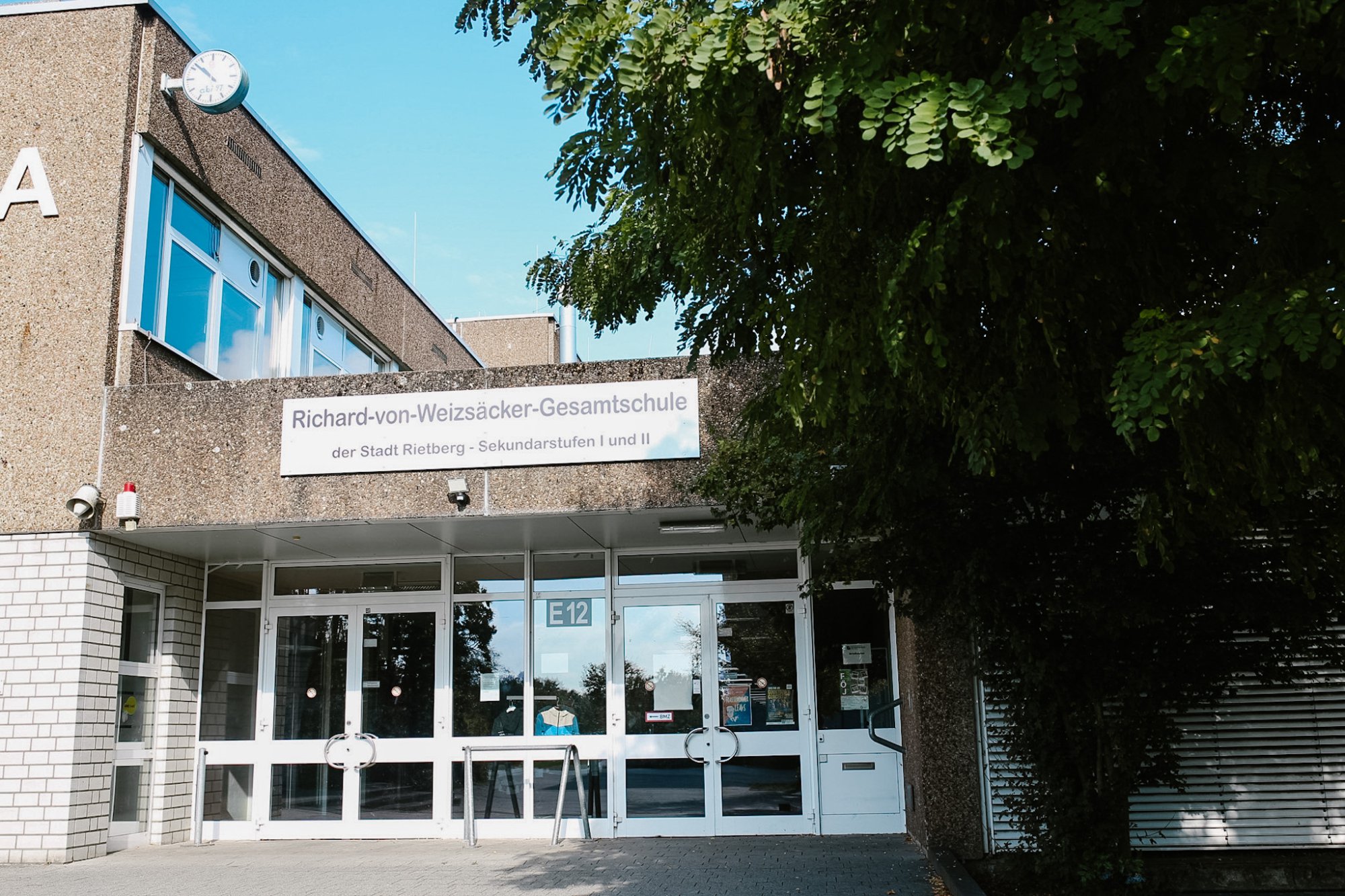 Haupteingang der Schule mit Glastüren und Vordach. Über dem Eingang steht auf einem Schild: Richard-von-Weizsäcker-Gesamtschule der Stadt Rietberg, Sekundarstufen I und II.