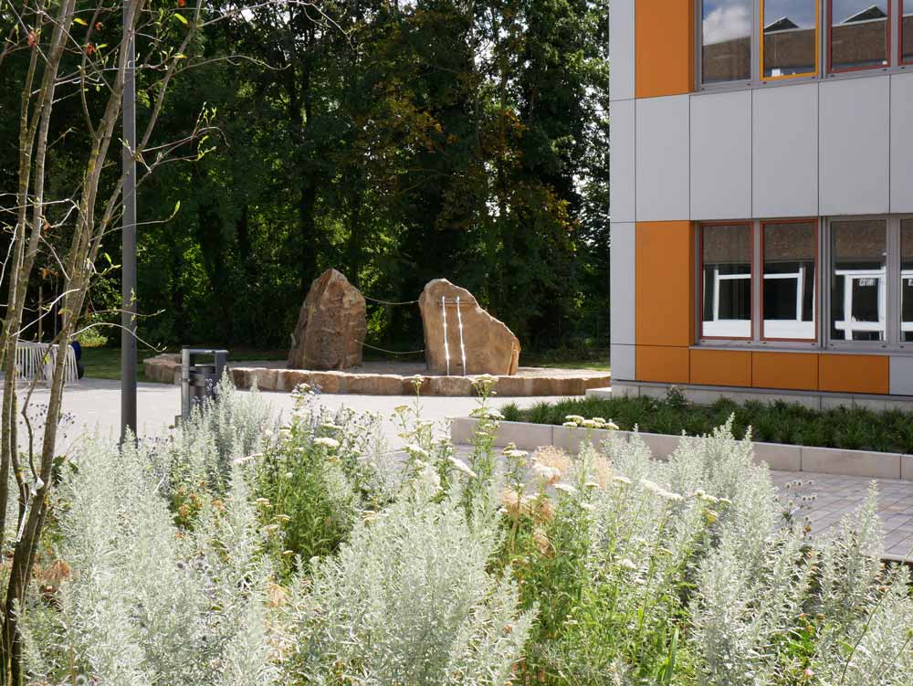Schulhof im Sommer mit blühenden Wildstauden im Vordergrund. Dahinter zwei große Natursteine auf einem runden Steinsockel und rechts das B-Gebäude mit orangefarbenen Fassadenelementen.