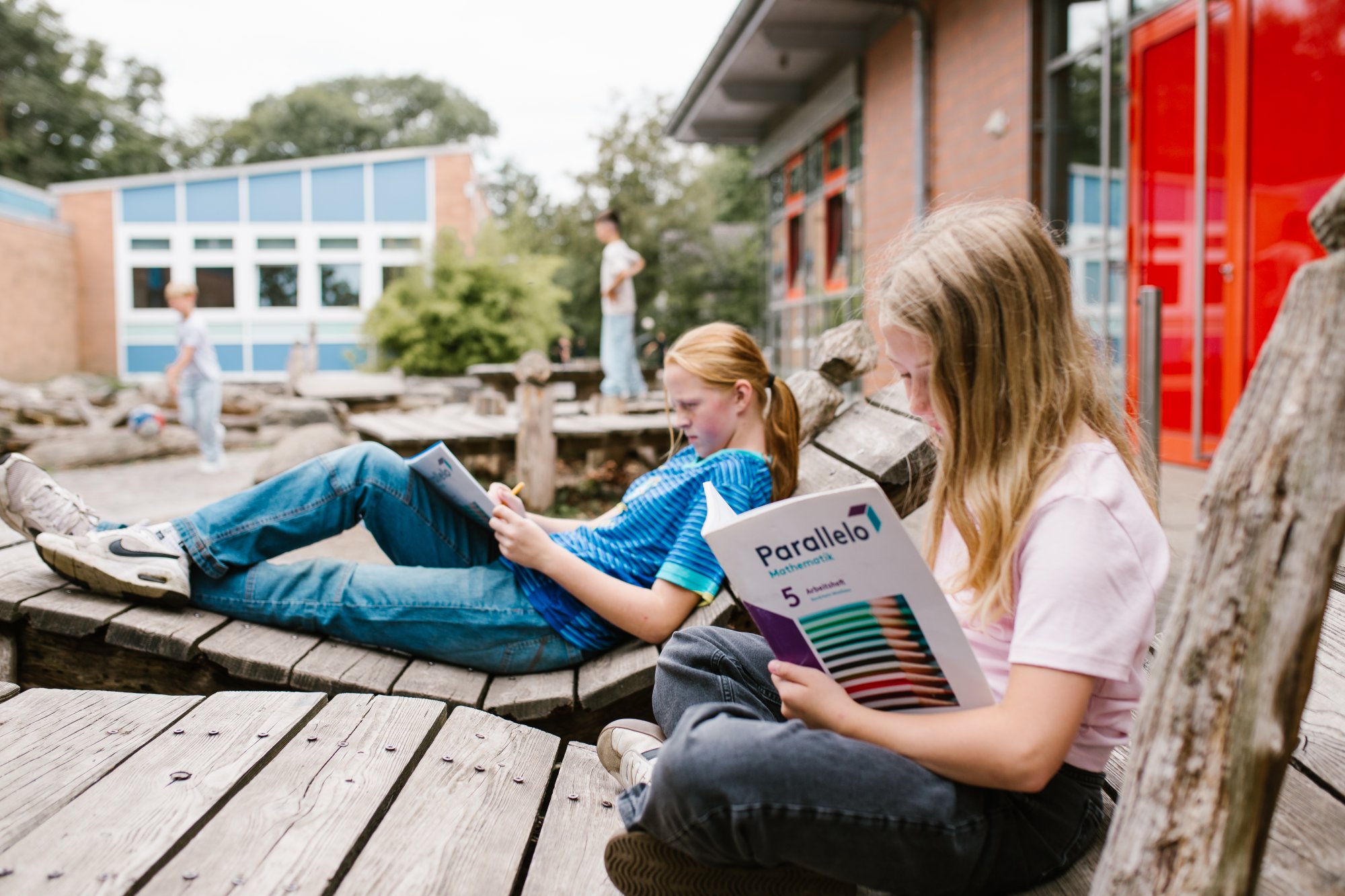 Zwei Kinder sitzen auf Holzbänken im Schulhof und arbeiten in einem Mathematik-Arbeitsheft. Im Hintergrund das Schulgebäude mit blauen und roten Fassadenelementen.