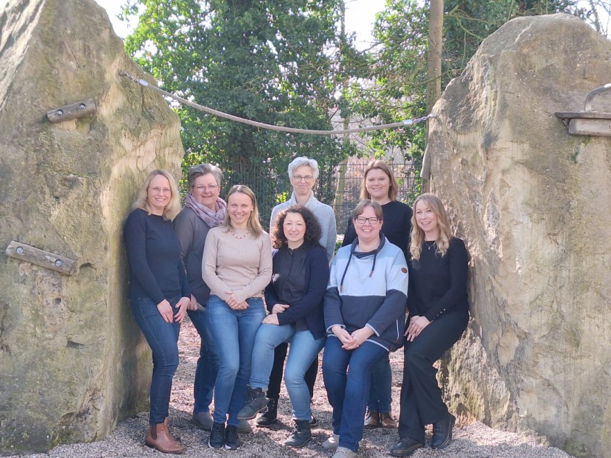 Acht Frauen aus dem Vorstand des Fördervereins stehen und sitzen zwischen zwei großen Kletterfelsen auf dem Schulhof.
