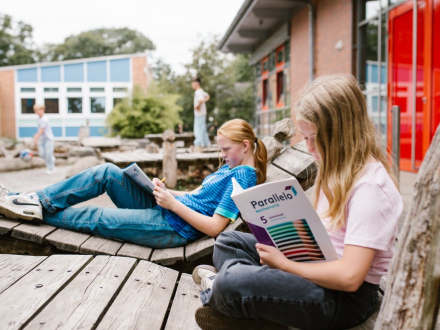 Zwei Kinder sitzen auf Holzbänken im Schulhof und arbeiten in einem Arbeitsheft, im Hintergrund das Schulgebäude